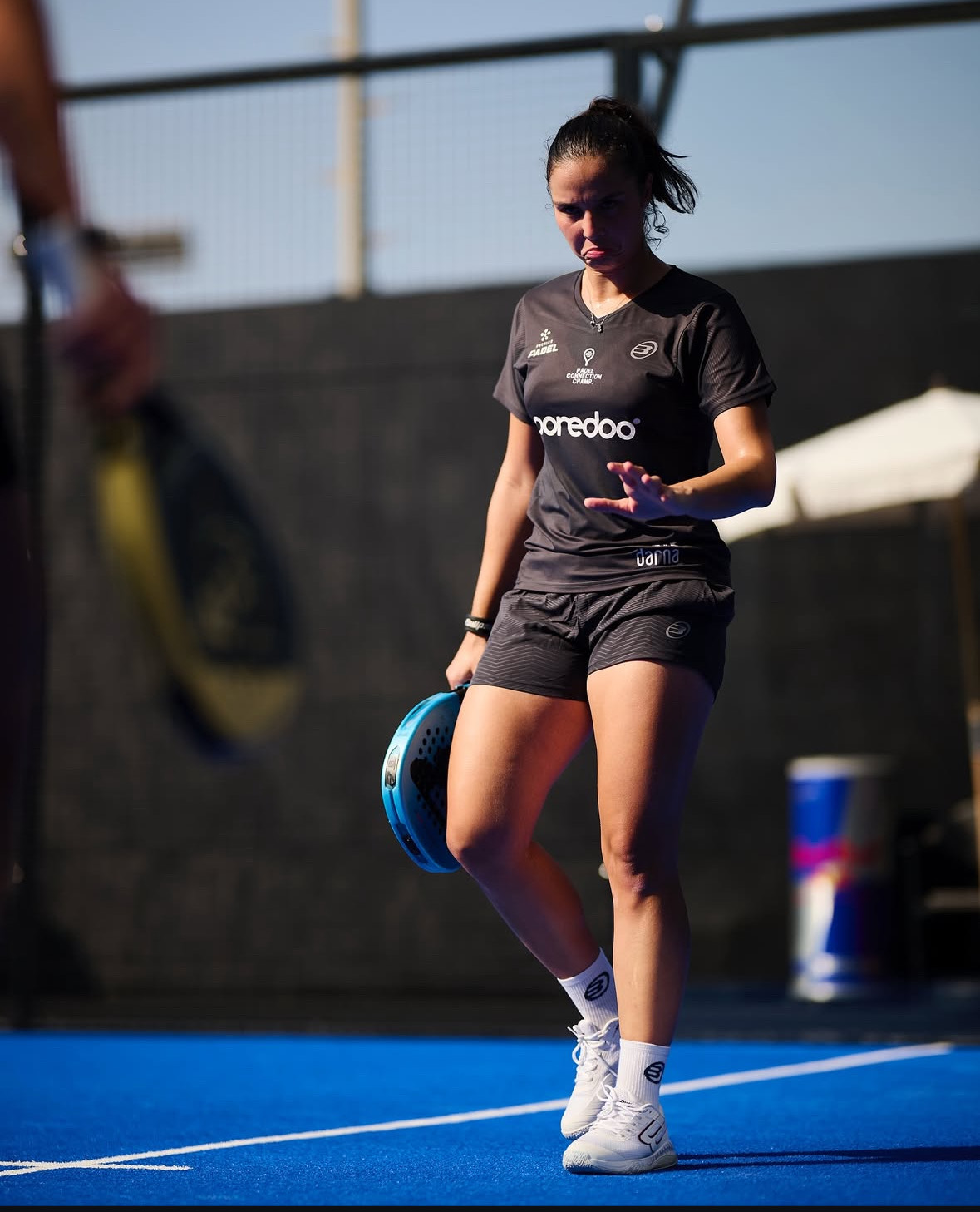 Dora Chamli walking between points on an outdoor blue court, racket in hand