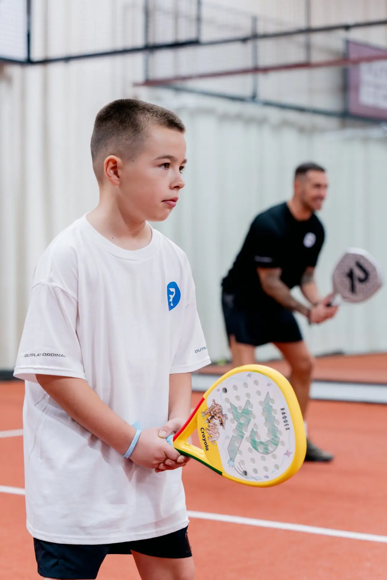 A boy waits for the ball on an indoor padel court holding a yellow Raquex x Crayola racket, with a coach in the background