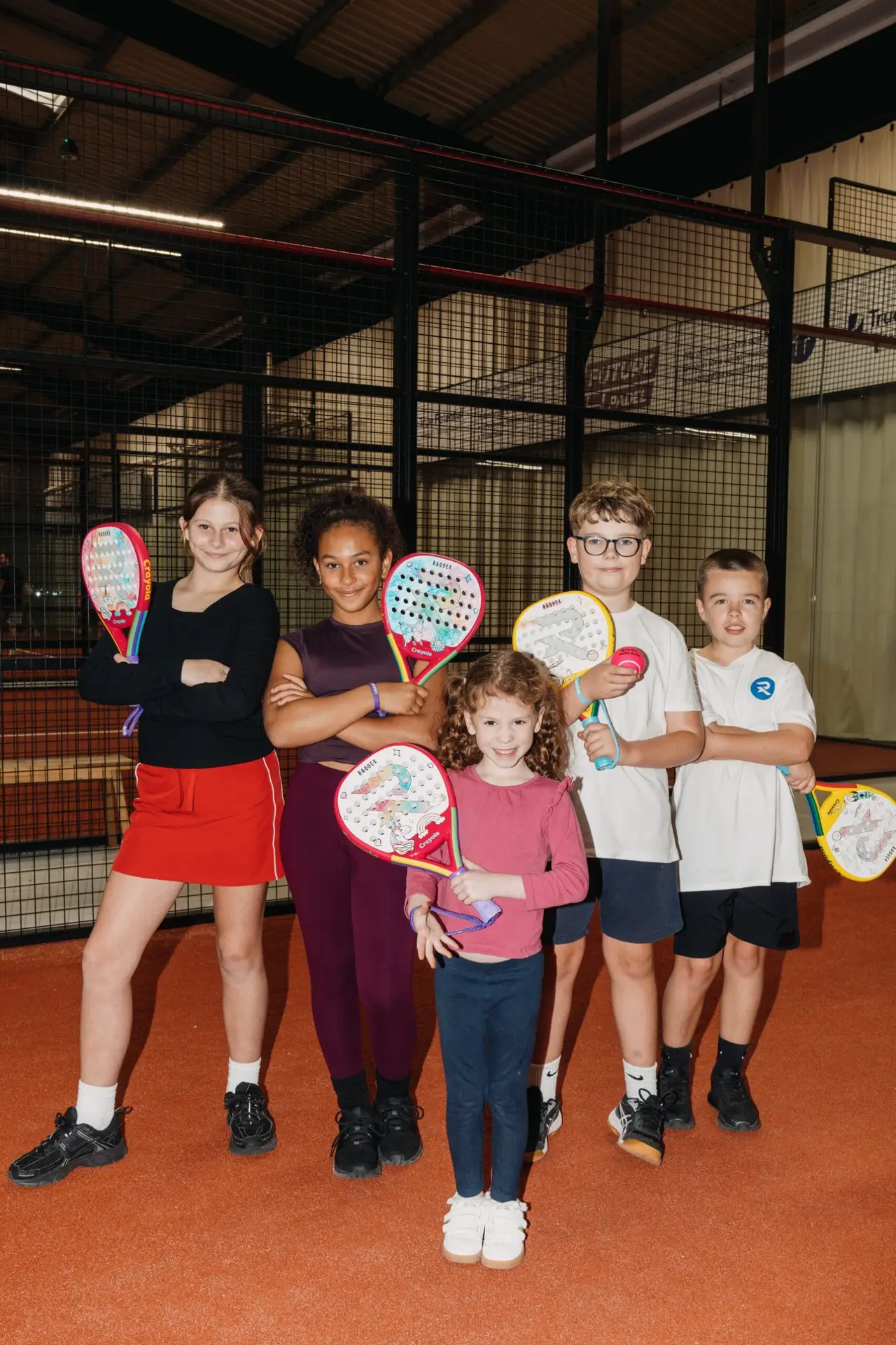 Five children stand on an indoor padel court holding Raquex x Crayola junior rackets